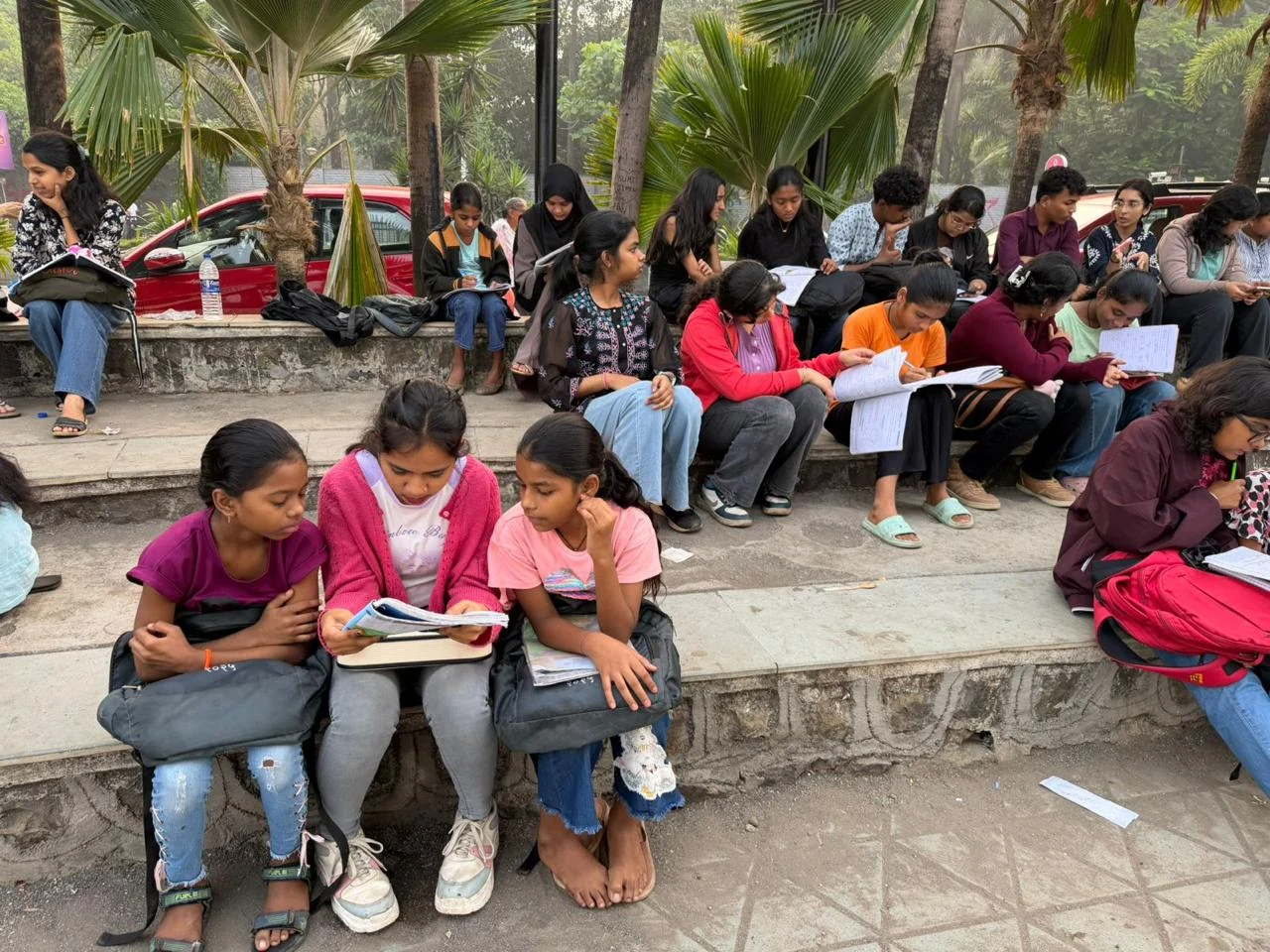 Children writing in notebooks beside volunteers, seated beneath palm trees